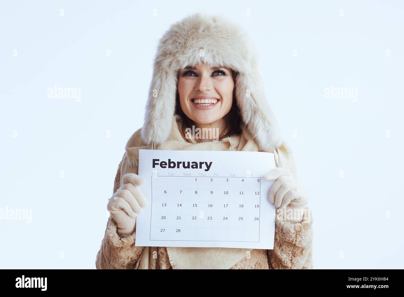 femme élégante heureuse en manteau d'hiver et chapeau de fourrure isolé sur blanc dans des gants blancs avec calendrier de février. Banque D'Images