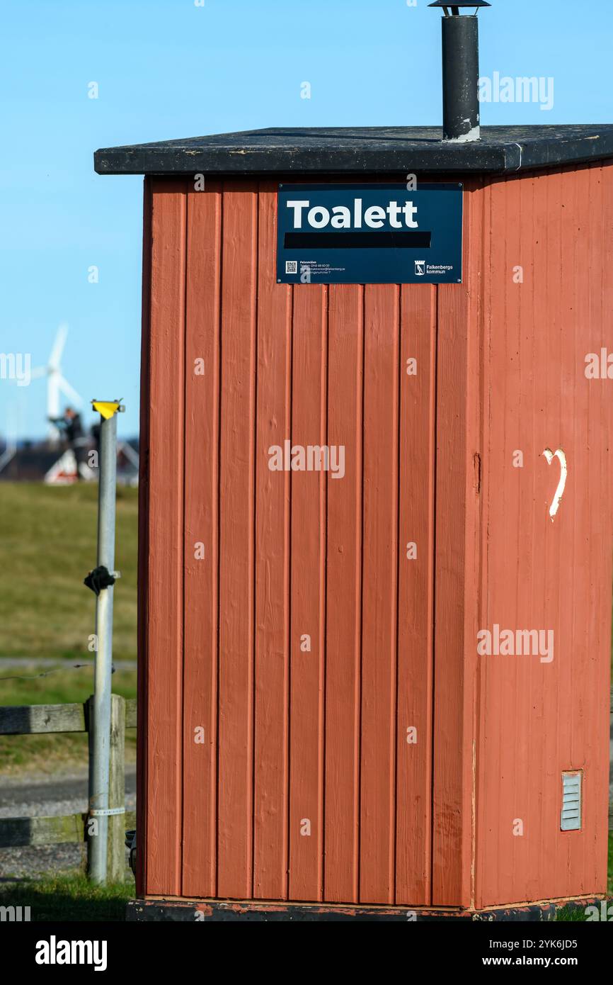 Une cabine de toilettes en bois rouge se dresse dans un champ, entouré de verdure. Les éoliennes peuvent être vues au loin contre un ciel bleu vif Banque D'Images