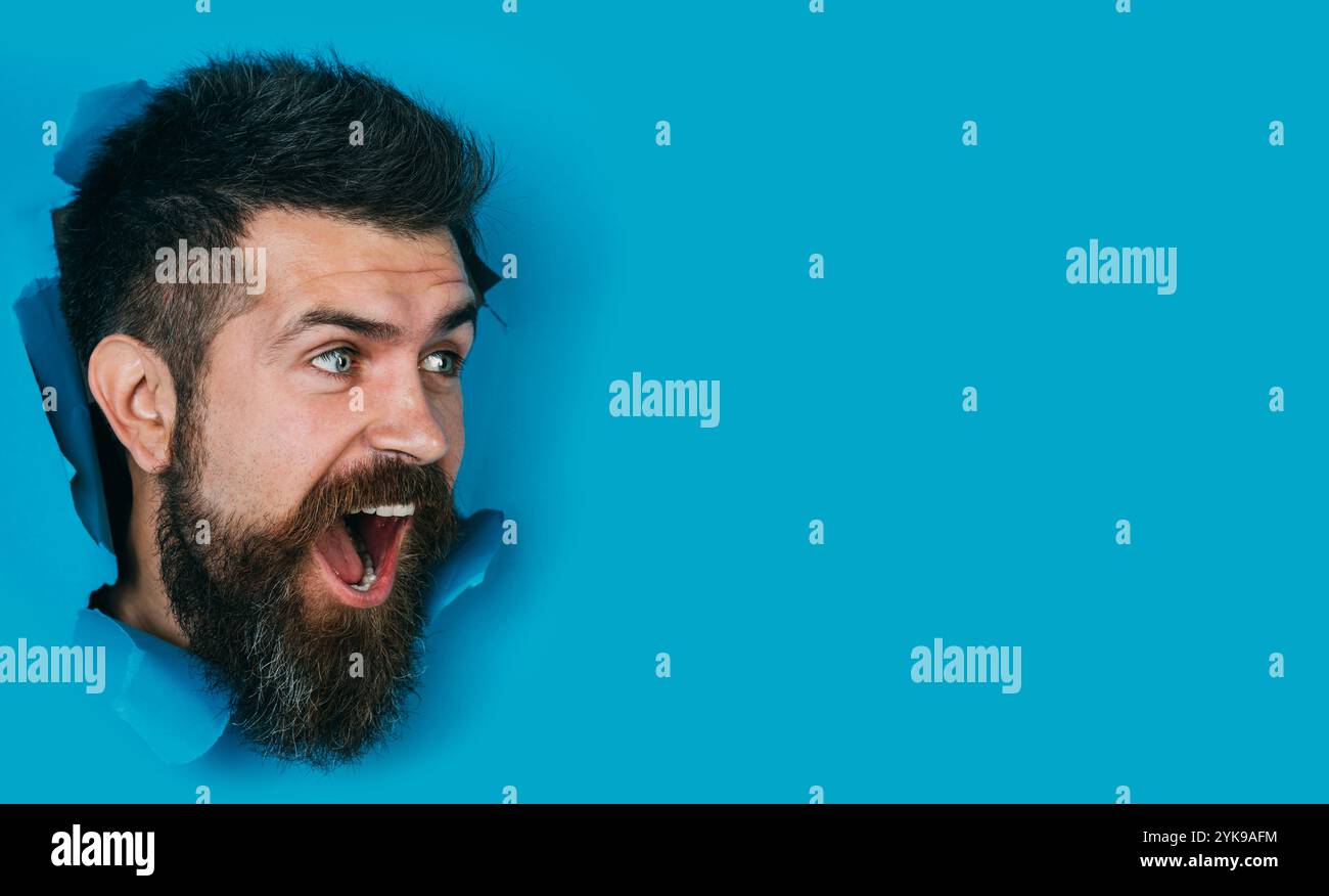Heureux homme barbu regardant à travers le trou dans le papier. Beau gars avec coiffure élégante faisant trou dans le mur de papier. Tête mâle regardant à travers le trou. Vente, Banque D'Images