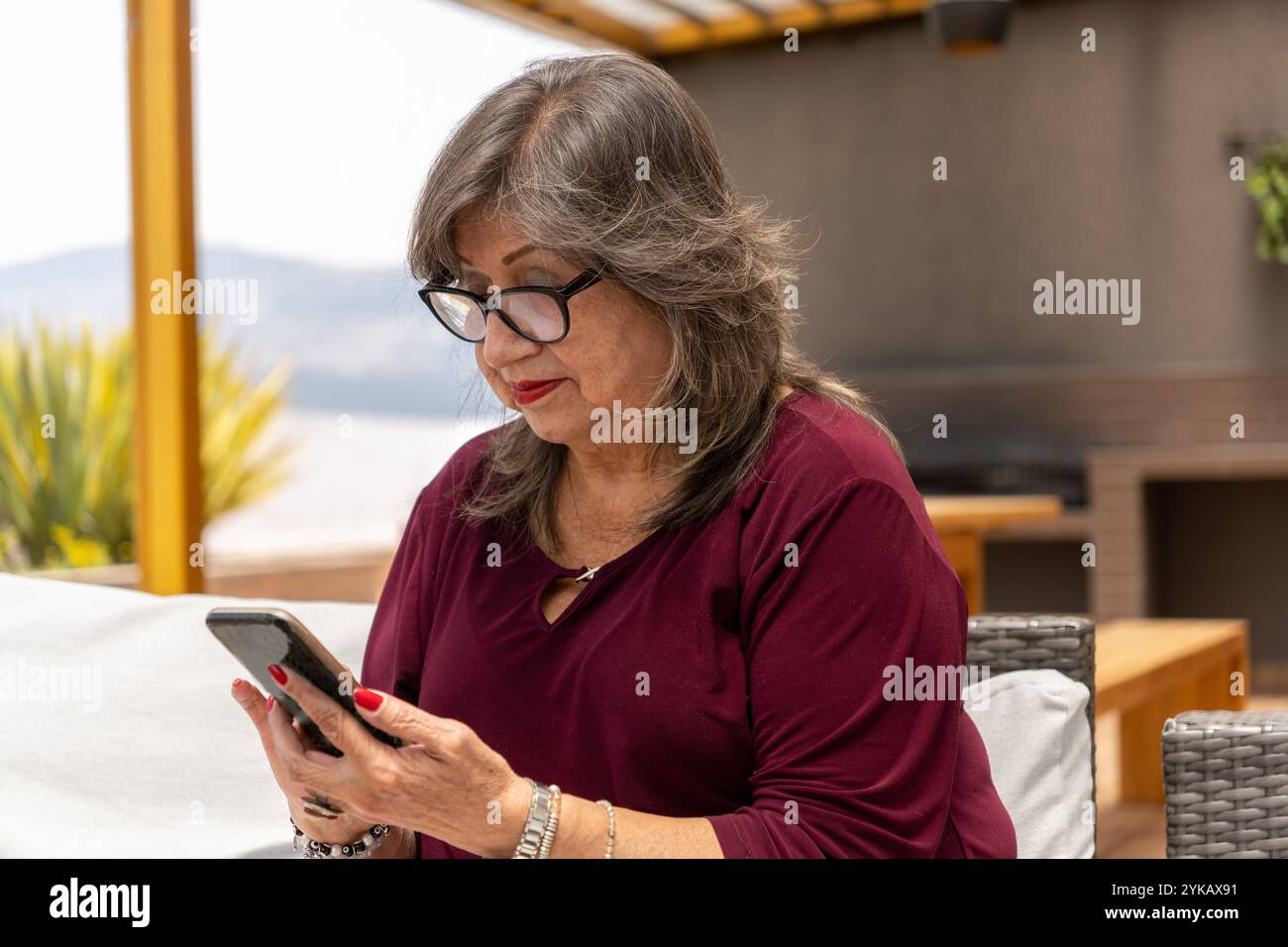 Femme Latina senior naviguant sur les médias sociaux sur son téléphone portable assis sur une terrasse extérieure le matin Banque D'Images