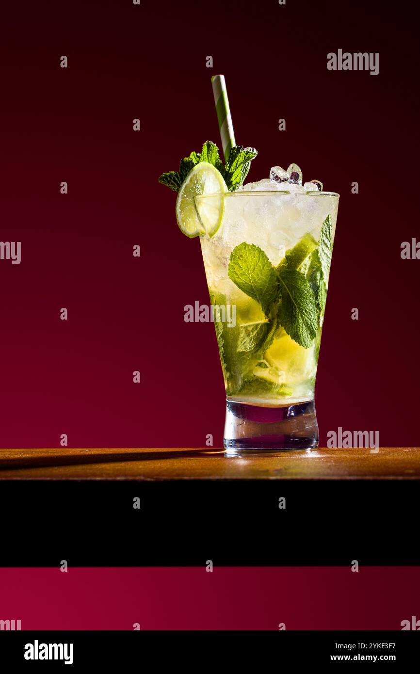 Un cocktail mojito vibrant garni de menthe fraîche et d'une tranche de citron vert dans un grand verre la boisson est sertie sur un fond rouge riche, mettant en valeur son Banque D'Images