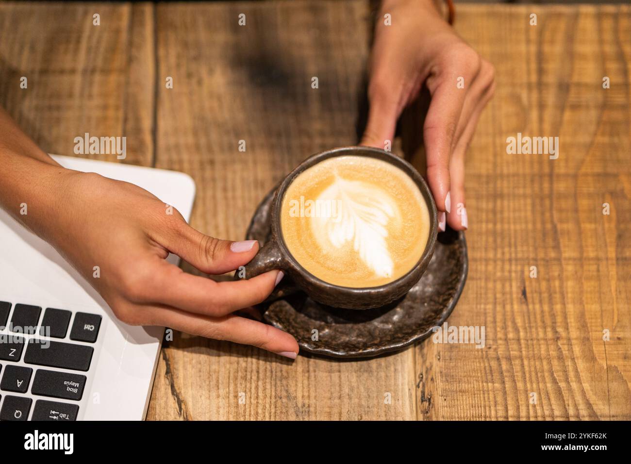 Une image en gros plan capturant les mains d'une femme d'affaires tenant un latte crémeux à côté de son ordinateur portable sur une table en bois rustique Banque D'Images