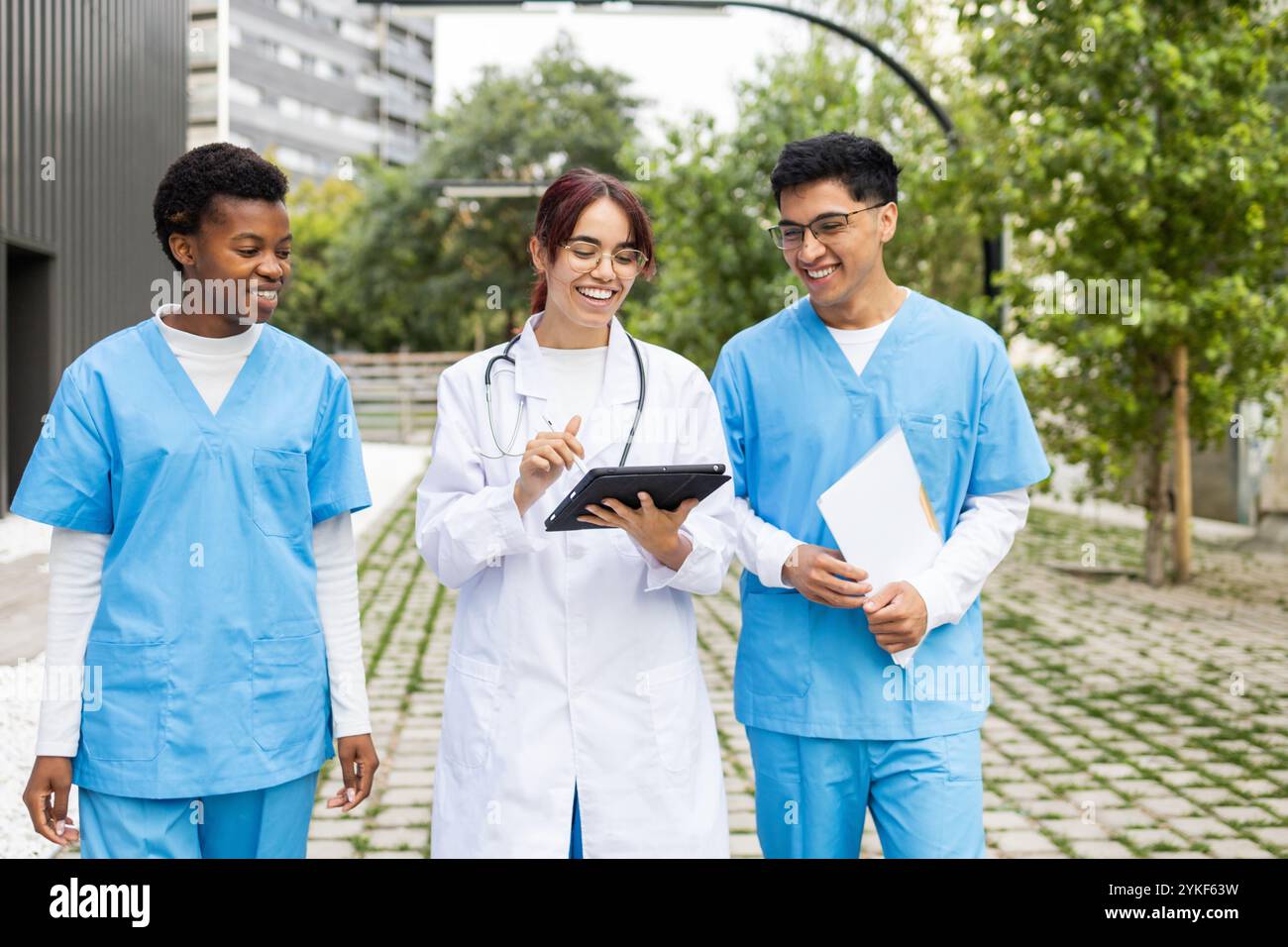 Trois jeunes étudiants en soins infirmiers de divers horizons conversent joyeusement tout en utilisant une tablette numérique en dehors de leur campus médical Banque D'Images