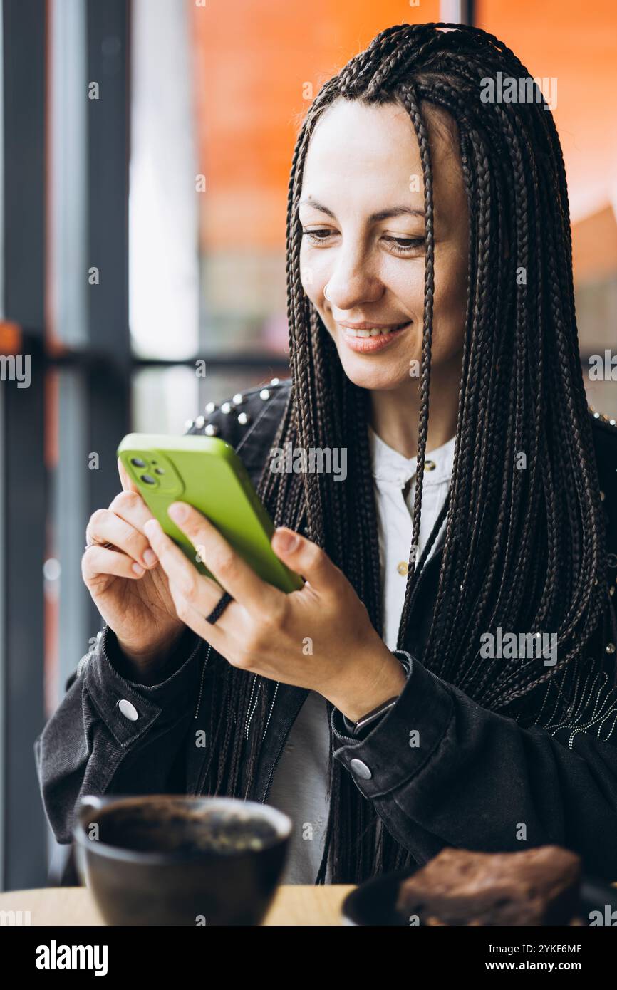 Une femme caucasienne élégante avec des tresses sourit tout en utilisant un smartphone et en buvant du café dans un café. Elle porte une veste noire et est fiancée avec Banque D'Images
