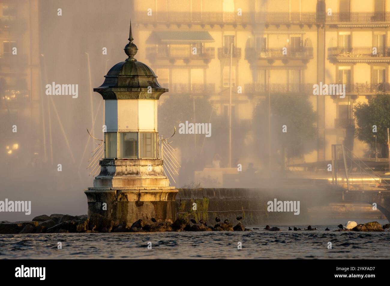 Une scène sereine à Genève capture un phare debout près de l'eau au lever du soleil, enveloppé dans une brume légère, avec la silhouette faible de construction urbaine Banque D'Images