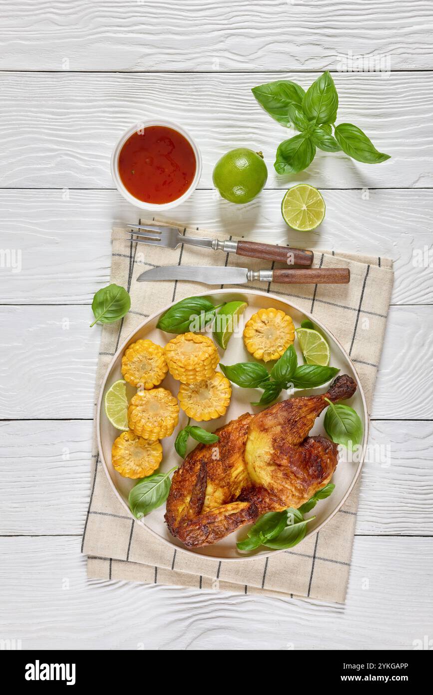 la moitié du poulet bbq avec des pièces d'épis de maïs et des feuilles de basilic frais sur l'assiette sur une table en bois blanc avec couverts, citron vert et sauce chili doux, vue verticale Banque D'Images