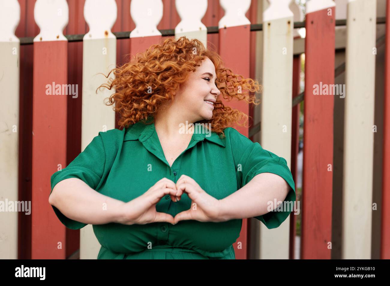 Portrait Happy plus Size femme avec cheveux rouges bouclés dans la danse de robe verte, faisant la forme de coeur avec les doigts à l'extérieur sur fond coloré lumineux Banque D'Images