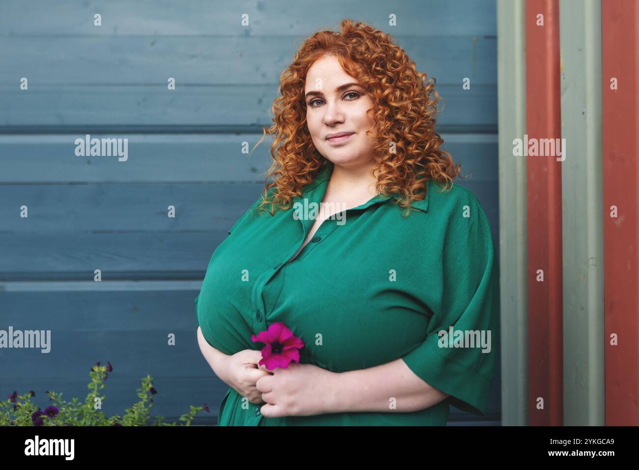 Femme de taille plus avec cheveux rouges bouclés dans des vêtements verts tient la fleur contre fond de mur bleu vibrant, beauté et confiance à l'extérieur Banque D'Images