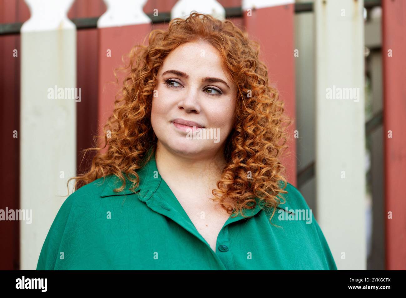 Portrait de belle femme grande taille avec des cheveux rouges bouclés dans des vêtements verts, regardant de côté sur fond rouge à l'extérieur en été Banque D'Images