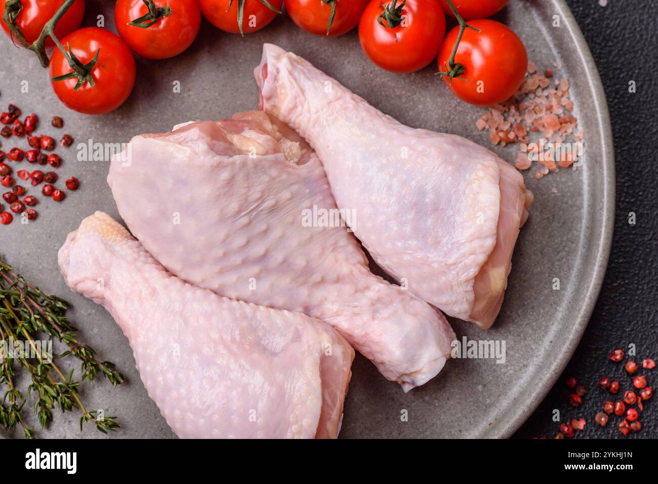 Cuisses de poulet crues avec du sel, des épices, des herbes et des tomates sur un fond de béton foncé Banque D'Images