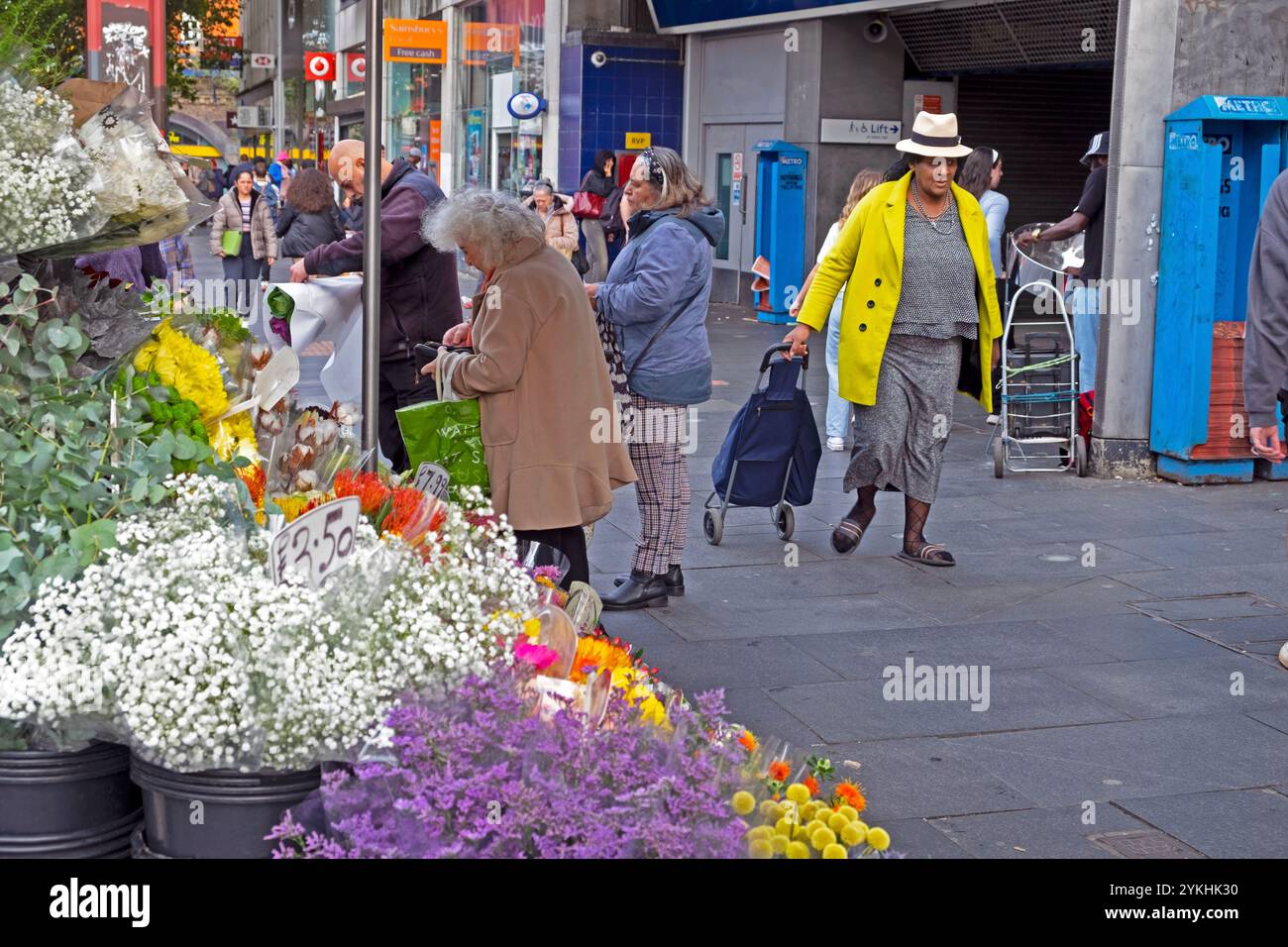 Chariot de magasinage senior plus âgé femme âgé portant chapeau de paille manteau jaune marchant dans la rue par l'étal de fleurs à Brixton Londres Angleterre KATHY DEWITT Banque D'Images
