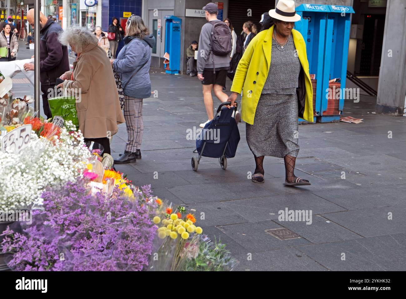 Chariot de magasinage senior plus âgé femme âgé portant chapeau de paille manteau jaune marchant dans la rue par l'étal de fleurs à Brixton Londres Angleterre KATHY DEWITT Banque D'Images