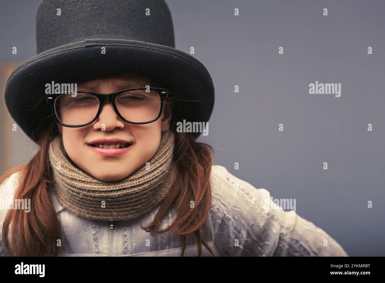 Une jeune fille de 8-9 ans joueuse dans un chapeau haut de gamme, des lunettes et une écharpe fait un visage drôle, rayonnant de bonheur et de créativité Banque D'Images