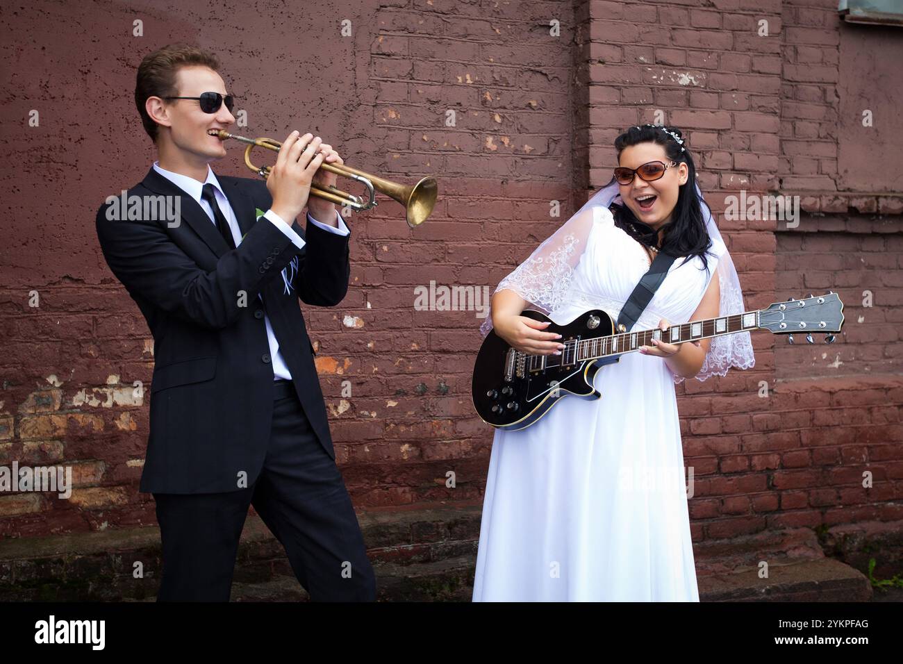 Le marié joue de la trompette et la mariée joue de la guitare lors d'une séance photo de mariage contre un mur de briques dans la rue Banque D'Images