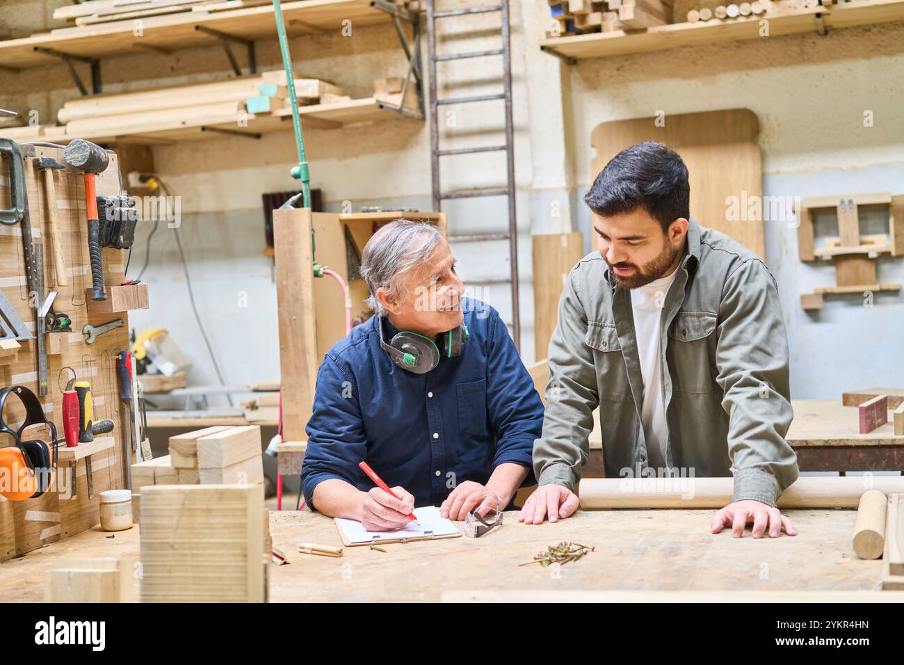 Un jeune apprenti reçoit les conseils d’un artisan expérimenté dans un atelier de menuiserie, mettant en valeur le travail d’équipe et le mentorat dans un set industriel Banque D'Images