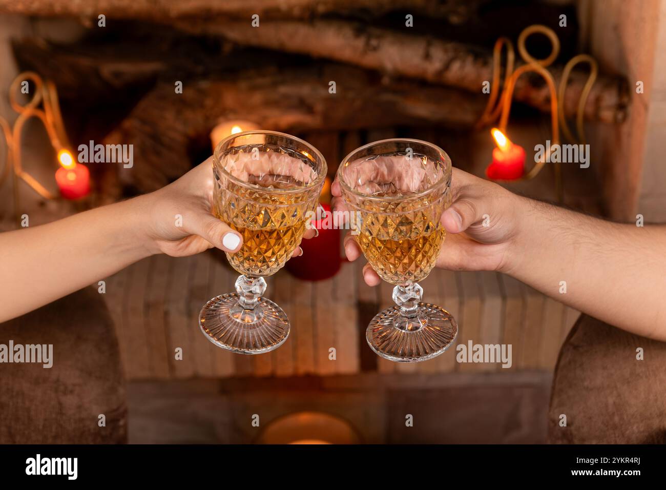 Une scène de soirée confortable avec deux mains levant des verres de cristal dans un toast près de la cheminée, entouré par la lueur chaude des bougies. Banque D'Images