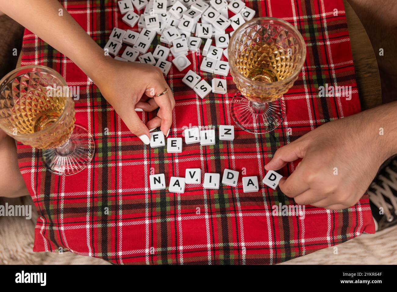 Une scène festive avec des mains arrangeant des tuiles orthographiant 'Feliz Navidad' sur une nappe à carreaux, avec des verres de vin. Noël et fête des fêtes Banque D'Images