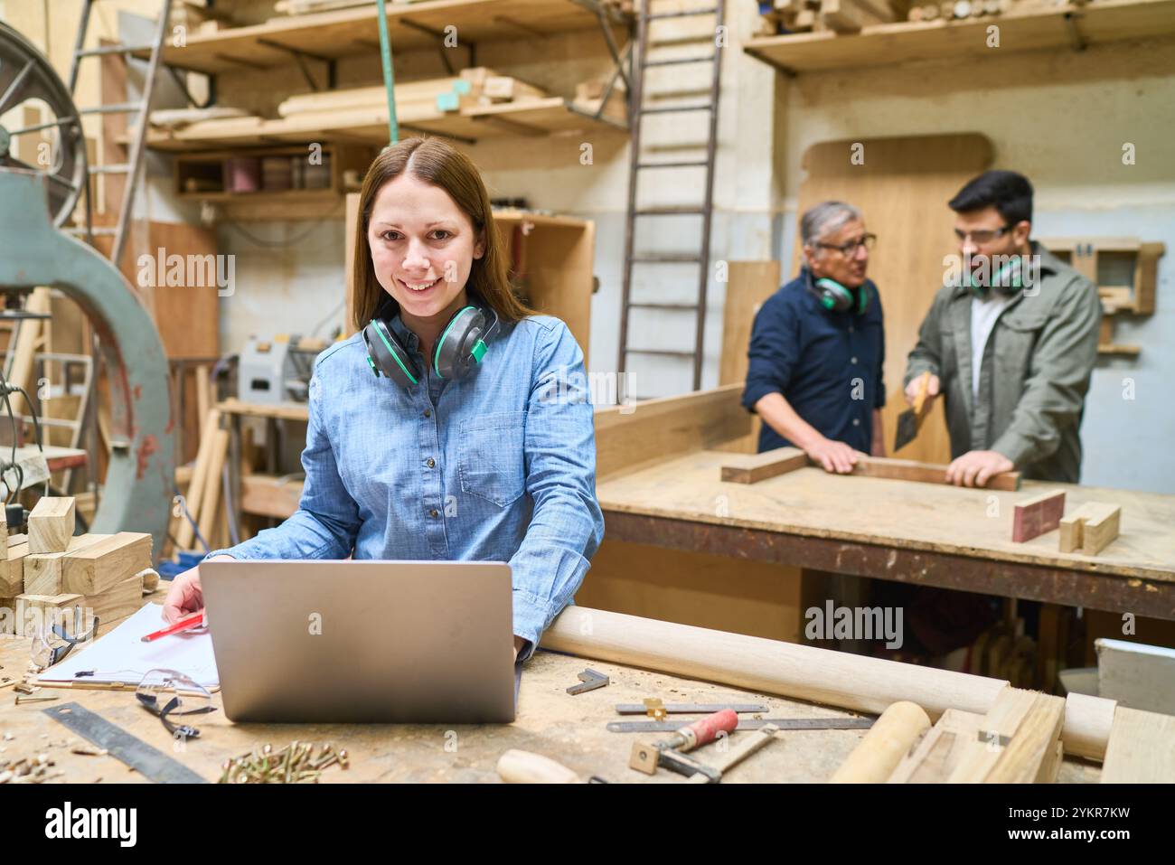 Une femme travaille sur un ordinateur portable dans un jardin forestier, avec des collègues collaborant à un projet de menuiserie en arrière-plan. La scène met en valeur le travail d'équipe Banque D'Images