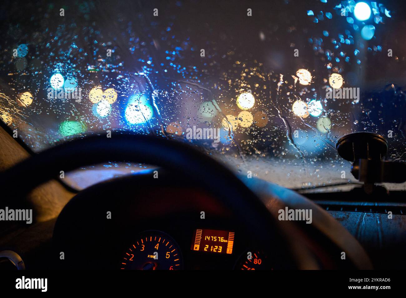 Gouttes de pluie sur un pare-brise de voiture avec des lumières de ville colorées floues la nuit, vues de l'intérieur. Un volant et un tableau de bord éclairés par une lumière douce Banque D'Images