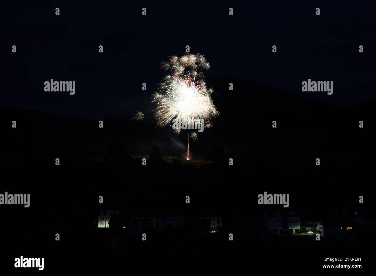 Une fusée de feu d'artifice dans le ciel sur une montagne la nuit, quelques maisons illuminées au premier plan Banque D'Images