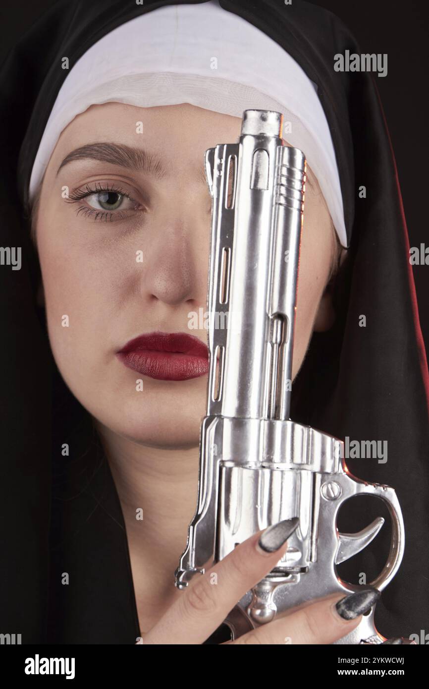 Portrait en gros plan d'une nonne tenant un revolver argenté. Jeune tueuse vêtue d'une robe de soeur religieuse Banque D'Images