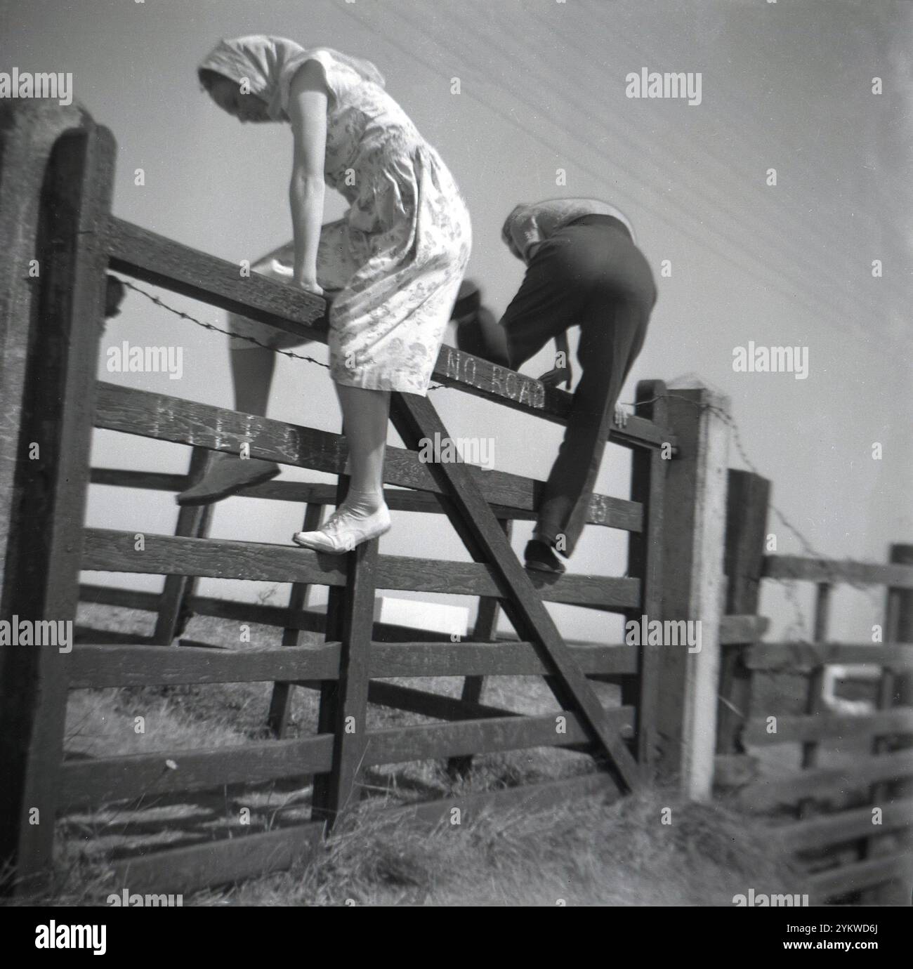 Années 1950, historique, un homme et une femme, qui porte une robe et une écharpe de cheveux, grimpant au-dessus d'un champ traditionnel en bois de 5 barres ou porte de ferme, Angleterre, Royaume-Uni. Banque D'Images