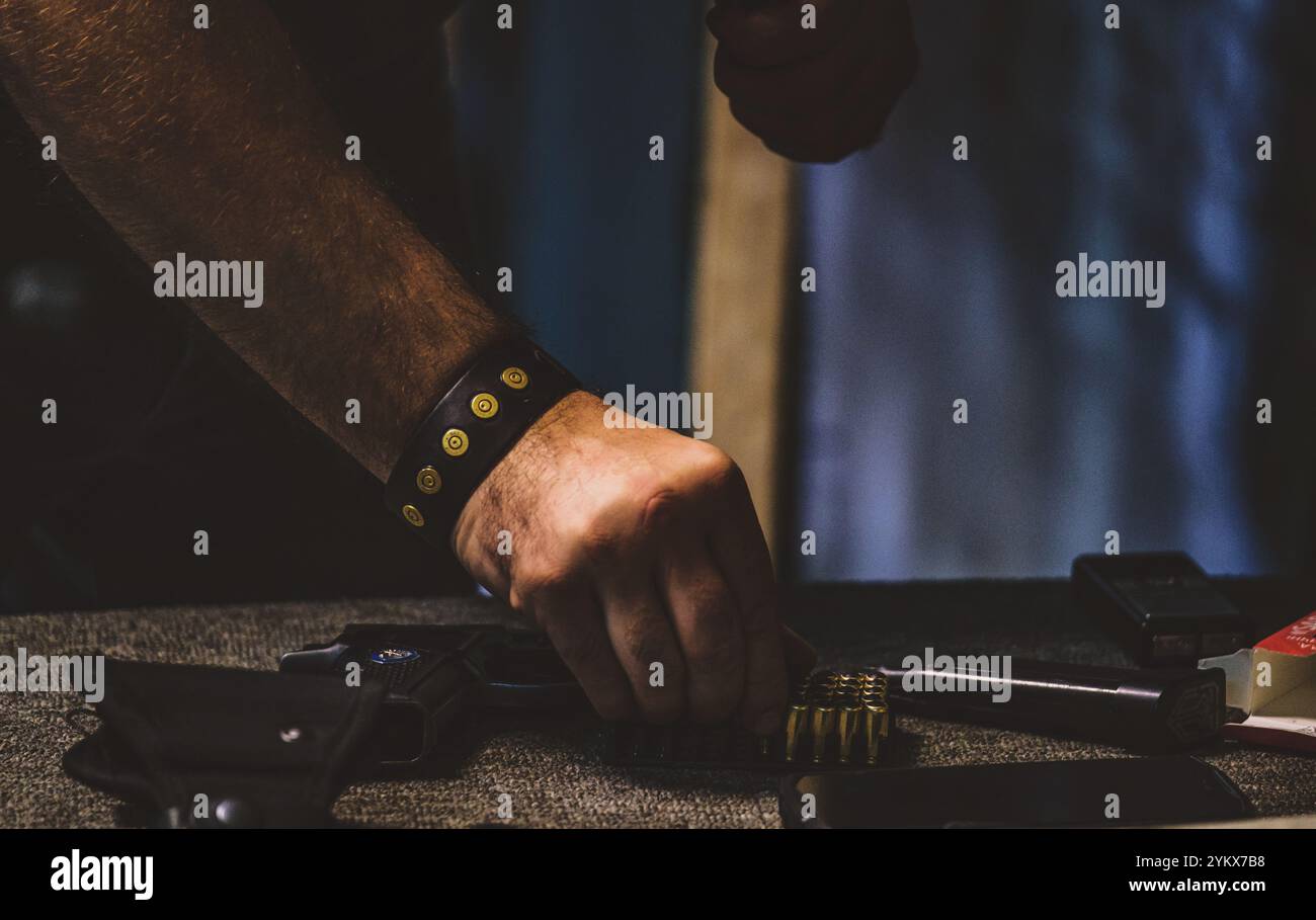 Homme manipulant des munitions. Sur la table, il y a une arme de poing dans un étui, une rangée de balles, et ce qui semble être un magazine ou un clip pour charger le Banque D'Images