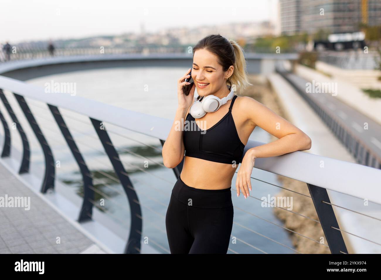 Une jogger s'arrête pour discuter sur son téléphone au bord de la rivière, portant un équipement d'entraînement et des écouteurs, embrassant le soleil chaud et l'atmosphère sereine arou Banque D'Images