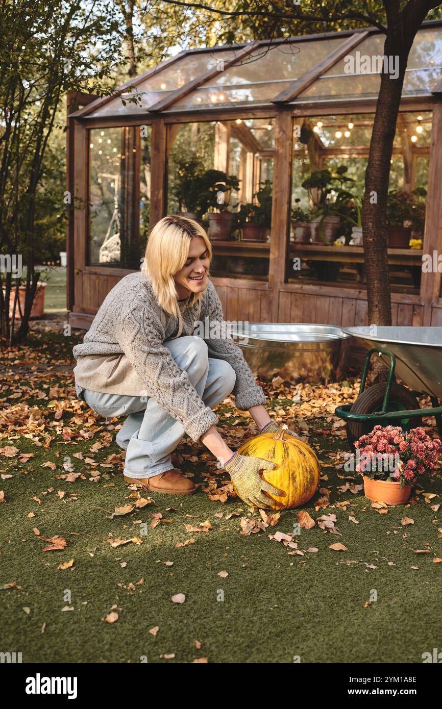 Un jeune homme beau aux cheveux blonds tend heureusement à une citrouille dans un jardin luxuriant rempli de feuilles d'automne. Banque D'Images