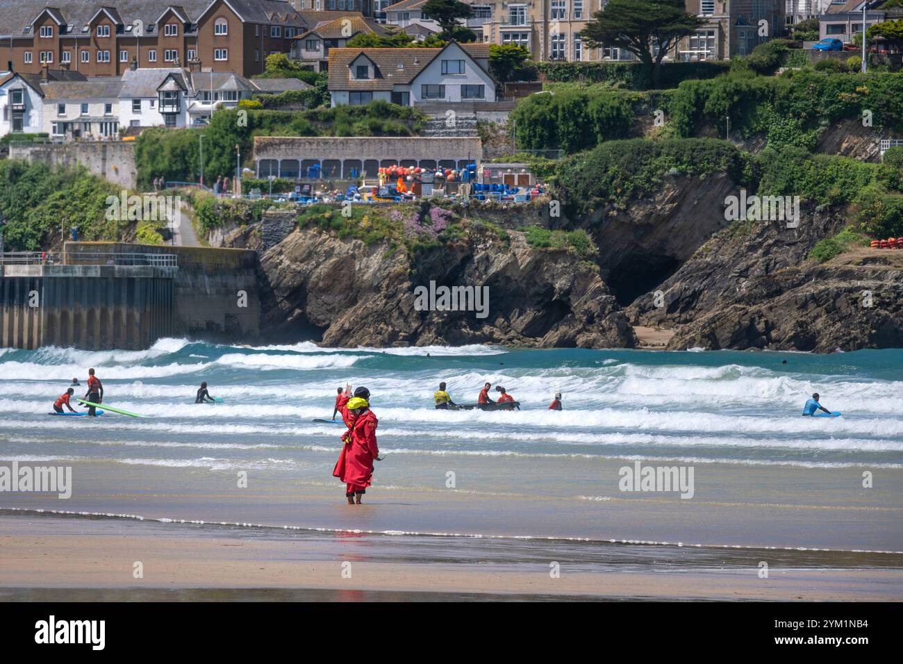 Un sauveteur de la RNLI Royal National Lifeboat institution en service debout sur le rivage regardant les surfeurs novices ayant une leçon de surf dans la mer à Towan Banque D'Images