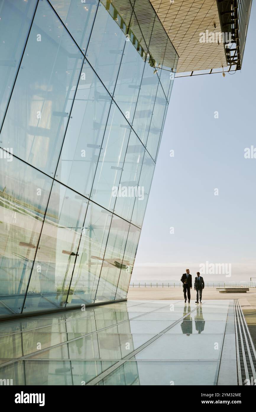 Façade en verre réfléchissant d'un bâtiment moderne projetant des motifs élégants sur le paysage urbain environnant montrant deux individus marchant le long de la surface miroir à la lumière du soleil Banque D'Images