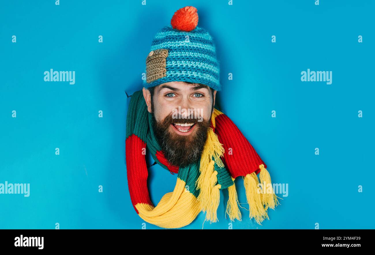 Homme barbu heureux en bonnet tricoté et écharpe regardant à travers le trou dans le papier. Beau mâle en bonnet tricoté et foulard coloré. Vêtements chauds tendance pour Banque D'Images
