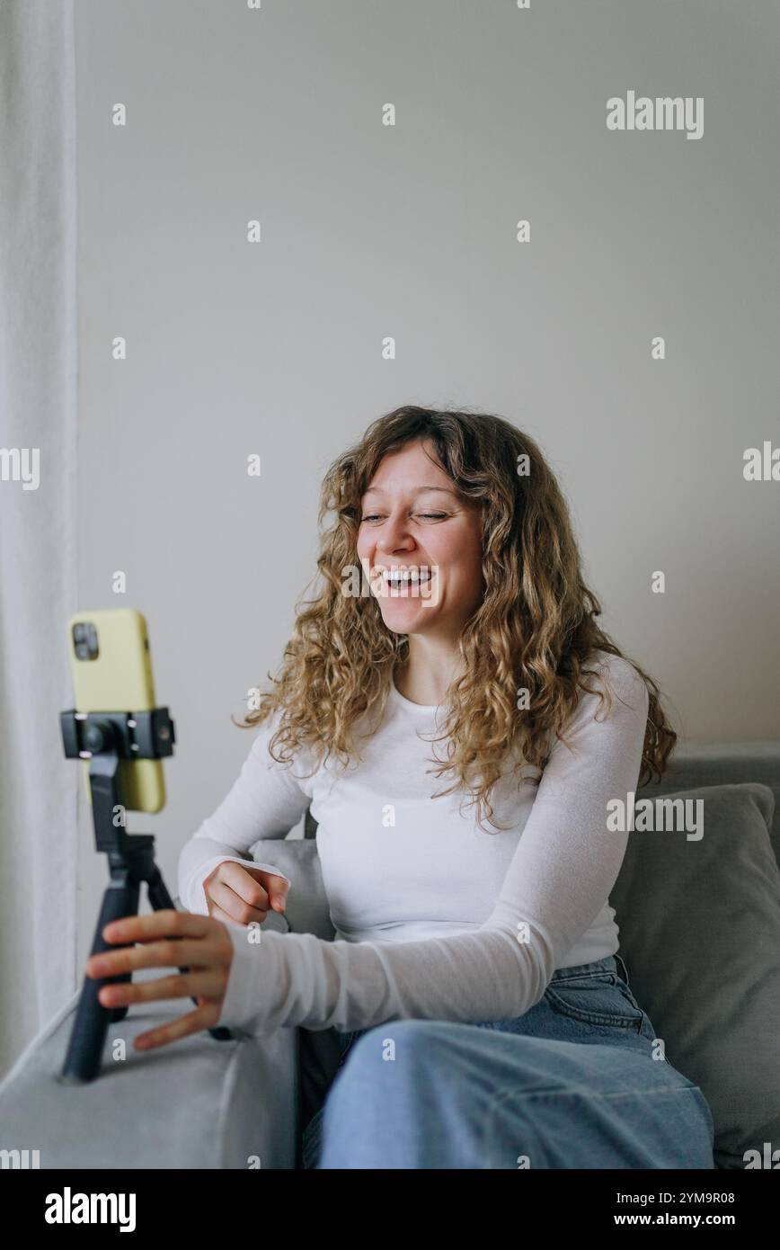 Jeune femme aux cheveux bouclés riant tout en enregistrant un blog vidéo à l'aide d'un smartphone sur un trépied, créant ainsi un contenu attrayant pour son public en ligne Banque D'Images