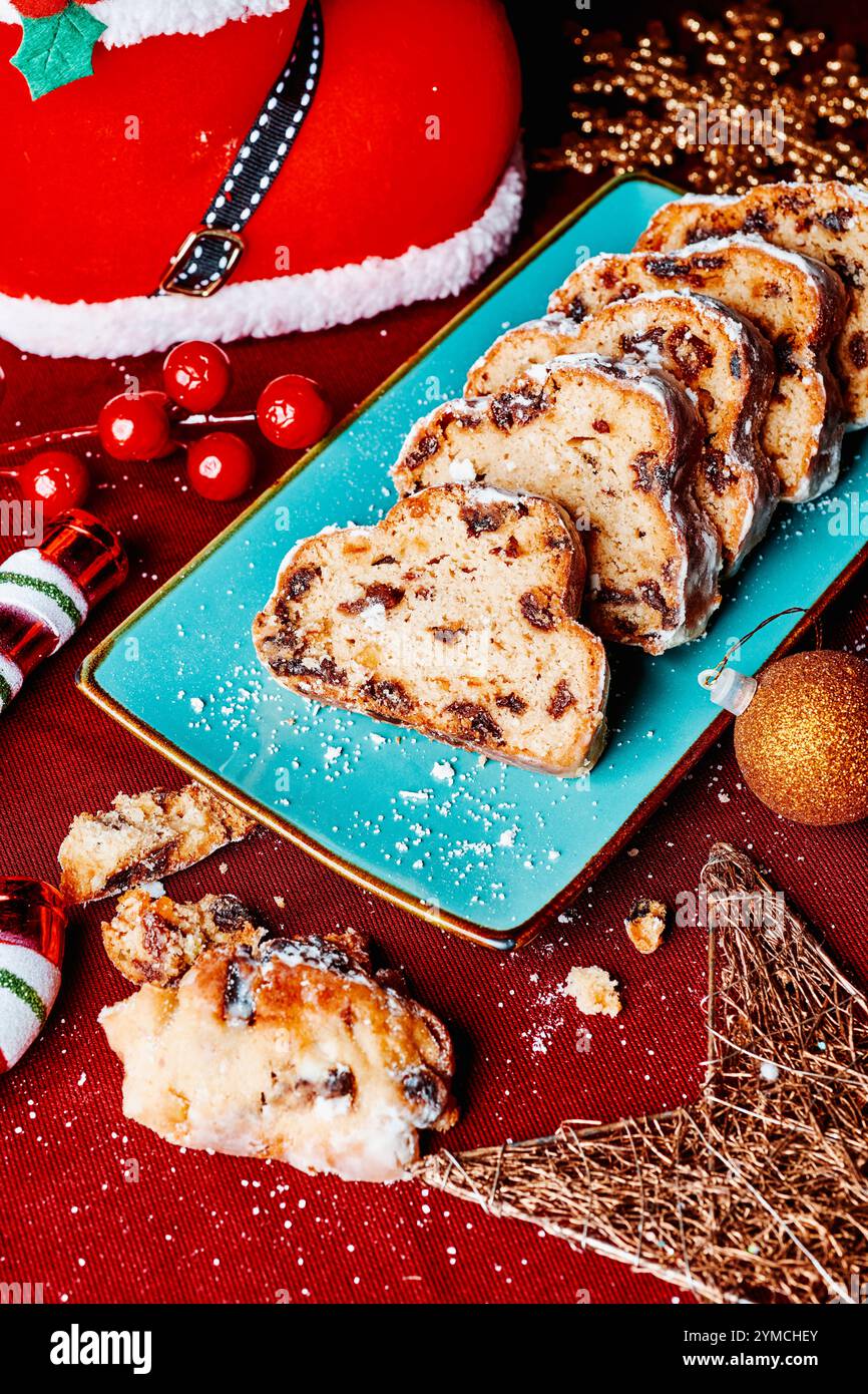 un stollen de noël tranché servi dans une assiette rectangulaire verte, sur une table avec une nappe rouge, à côté de quelques ornements de noël Banque D'Images