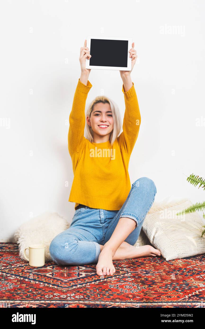 Femme caucasienne aux cheveux blonds dans un pull jaune tient une tablette blanche en altitude, assise les jambes croisées sur un tapis décoratif avec des éléments confortables. Banque D'Images