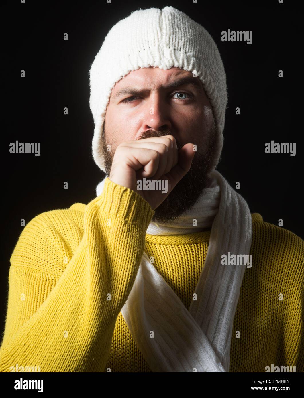 Portrait d'un malade barbu toussant dans son poing. Santé et médecine. Malade homme malade en pull jaune, écharpe blanche et chapeau toussant et Banque D'Images