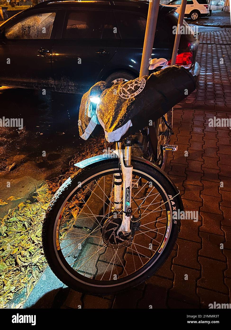 Kiev, Ukraine - 22 novembre 2024 : une bicyclette avec un équipement réfléchissant est garée dans une rue humide la nuit. Les feux incandescents améliorent la visibilité et la sécurité Banque D'Images