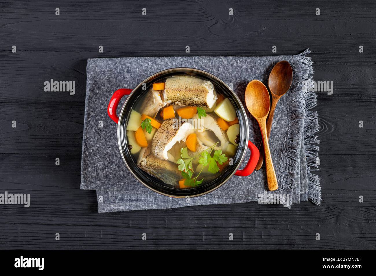 Soupe claire de brochet avec des légumes, du persil et des épices dans un pot rouge sur une table en bois noir avec des cuillères en bois, vue horizontale d'en haut, pose à plat Banque D'Images