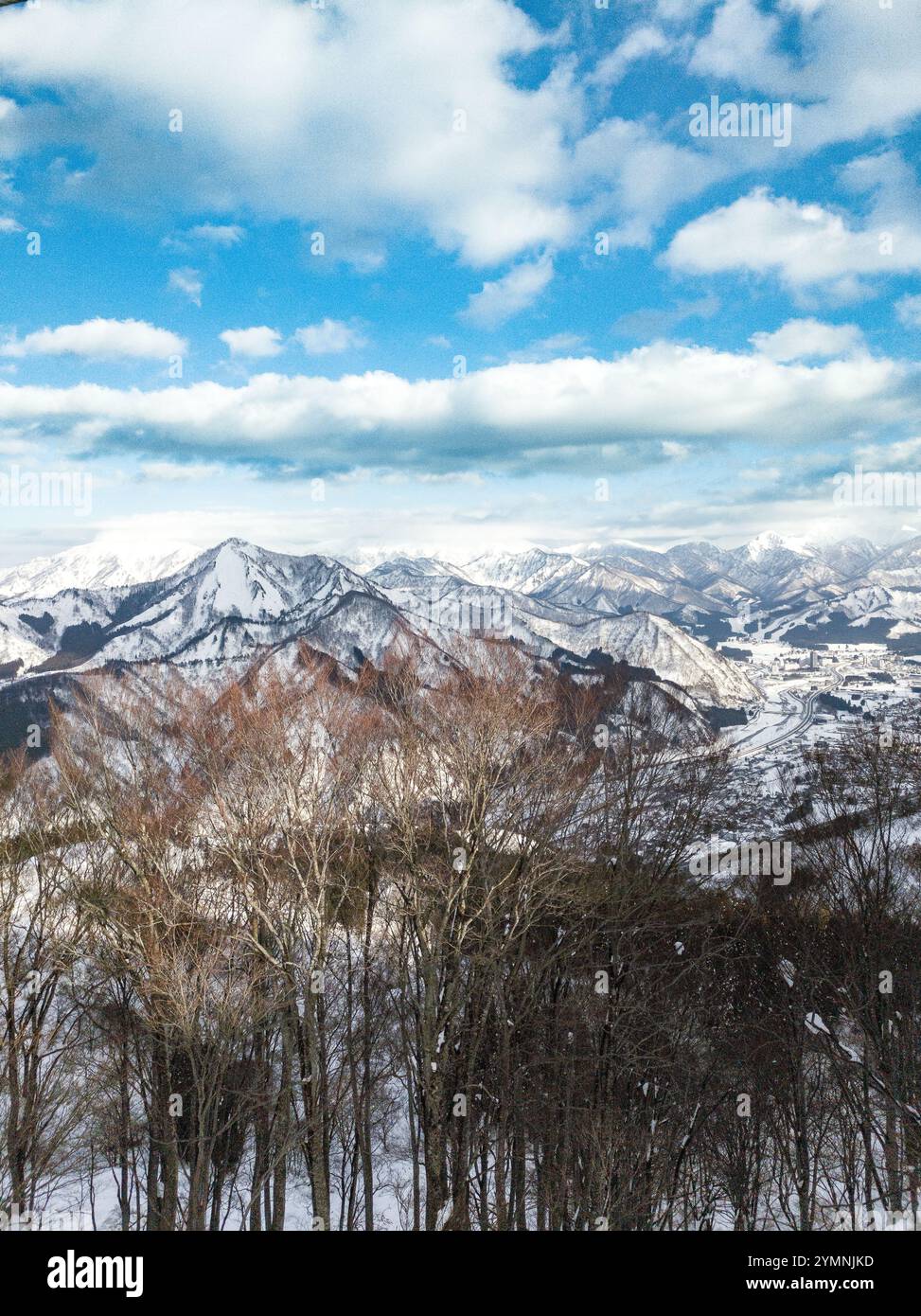 Montagnes japonaises enneigées sous un ciel bleu éclatant avec des nuages éparpillés, encadrées par des arbres d'hiver nus dans un panorama panoramique Banque D'Images