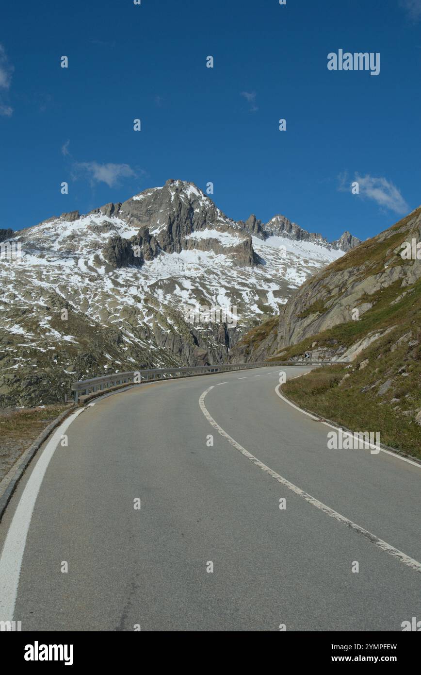 Route de montagne sinueuse dans les Alpes avec des sommets enneigés en arrière-plan sous un ciel bleu vif, idéal pour les voyages et les thèmes de la nature. Banque D'Images