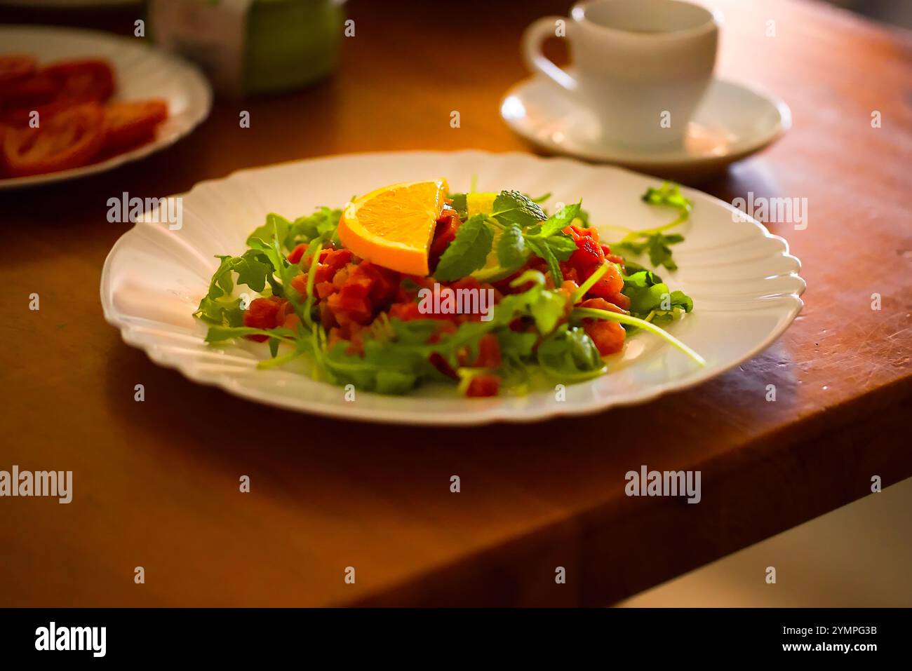 Assiette blanche contient tartare de thon avec roquette fraîche, menthe et tranches d'agrumes, sur une table en bois ; scène du petit déjeuner légèrement floue en arrière-plan. Banque D'Images