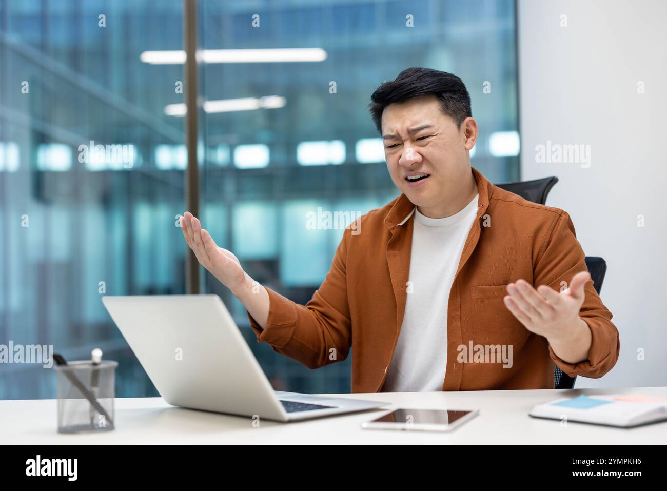 Homme d'affaires asiatique montrant la frustration tout en travaillant sur ordinateur portable dans le bureau. Exprimer le stress et la confusion avec les mains levées et l'expression déconcertée, téléphone sur le bureau. Banque D'Images