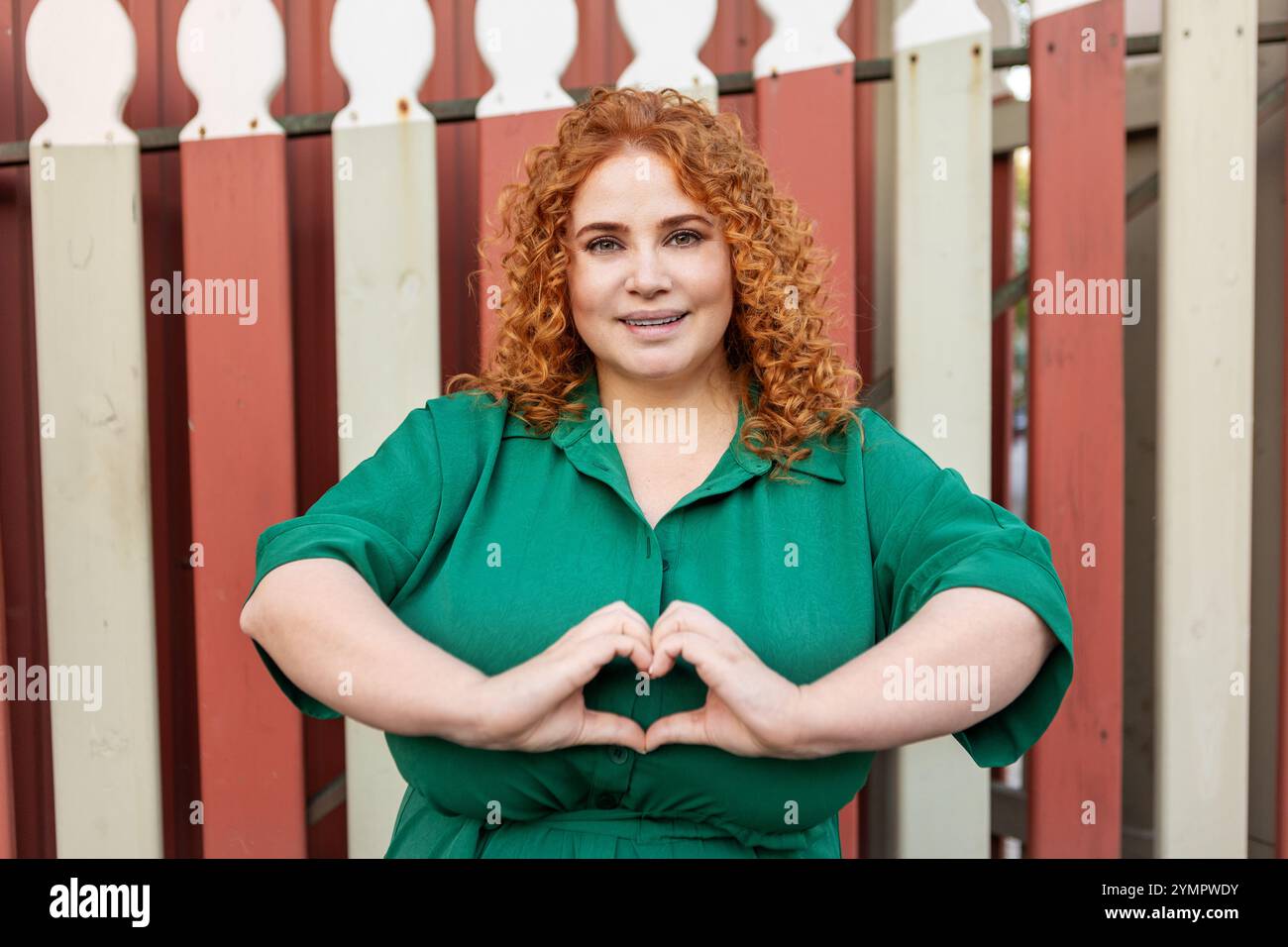 Bonne femme de taille plus souriante avec cheveux rouges bouclés en robe verte regardant la caméra, faisant geste de forme de coeur avec les doigts à l'extérieur contre Bright Co Banque D'Images