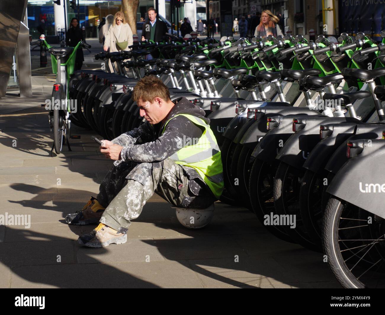 Constructeur de col bleu assis sur un casque de sécurité devant des vélos de location Lime tout en lisant un téléphone intelligent, Londres, Royaume-Uni Banque D'Images