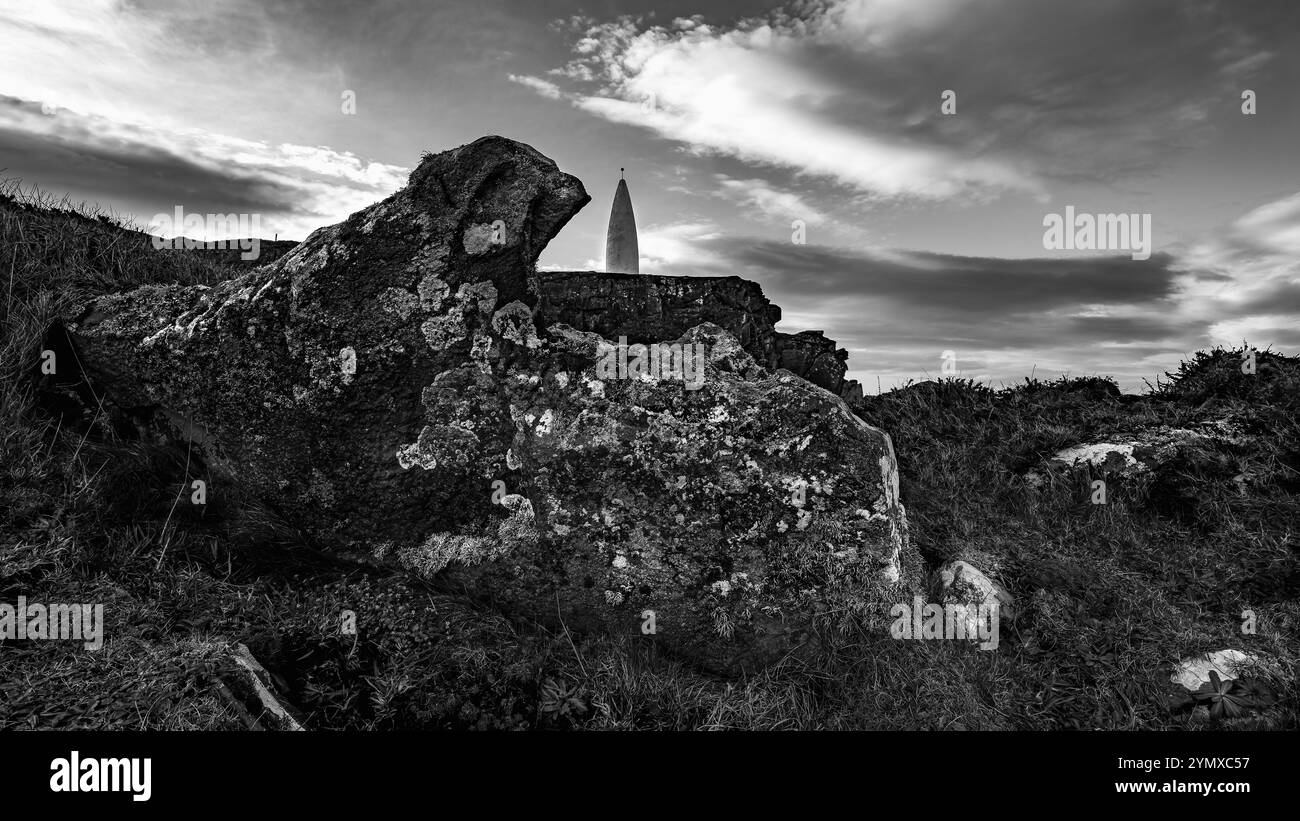 Vue d'une pente rocheuse avec des parcelles d'herbe et une structure haute, blanche et étroite au loin. Le ciel est généralement dégagé. Banque D'Images