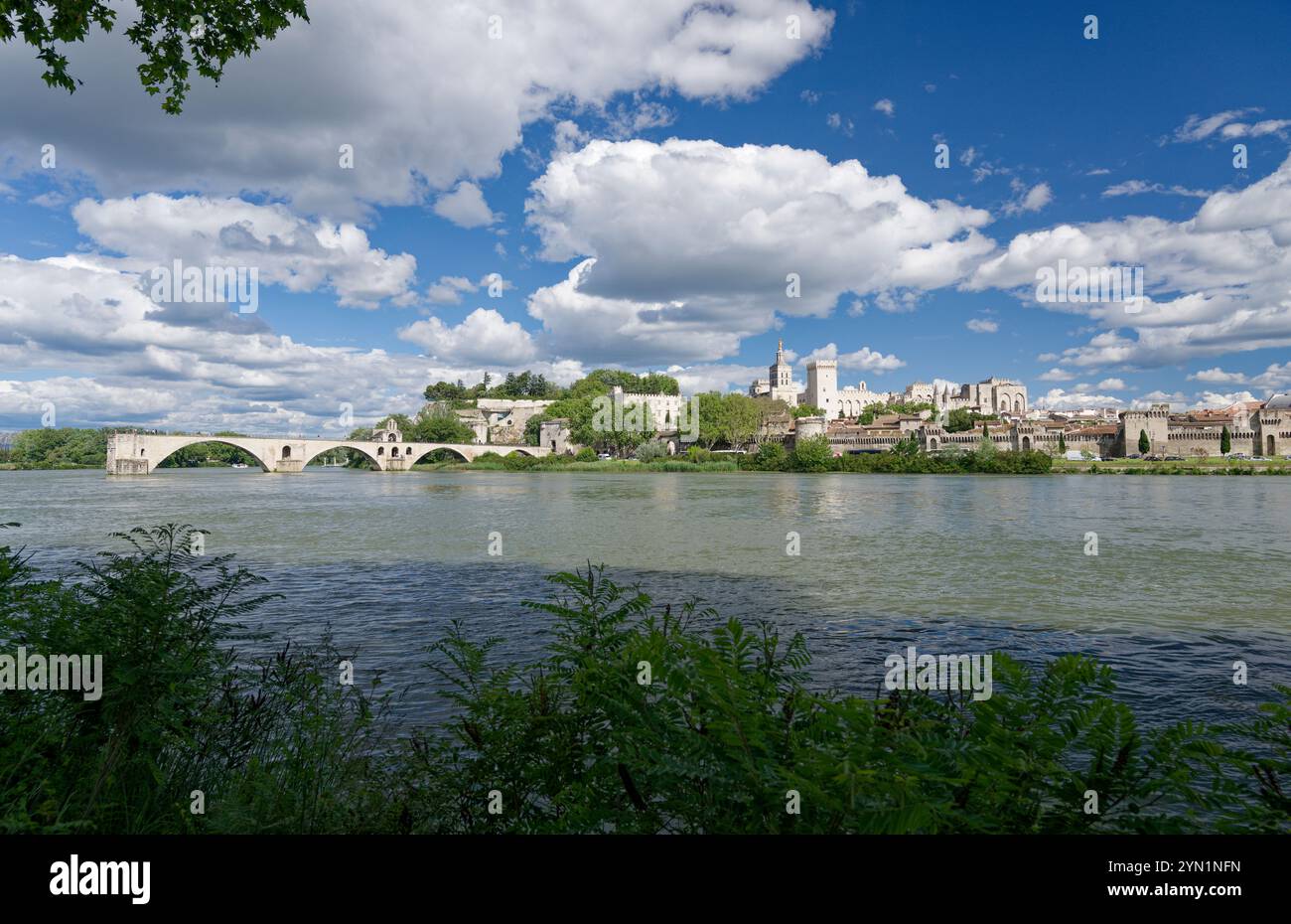 Paysage urbain historique d'Avignon avec le pont de pierre inachevé sur une rivière tranquille sous un ciel bleu vibrant avec des nuages moelleux. Banque D'Images