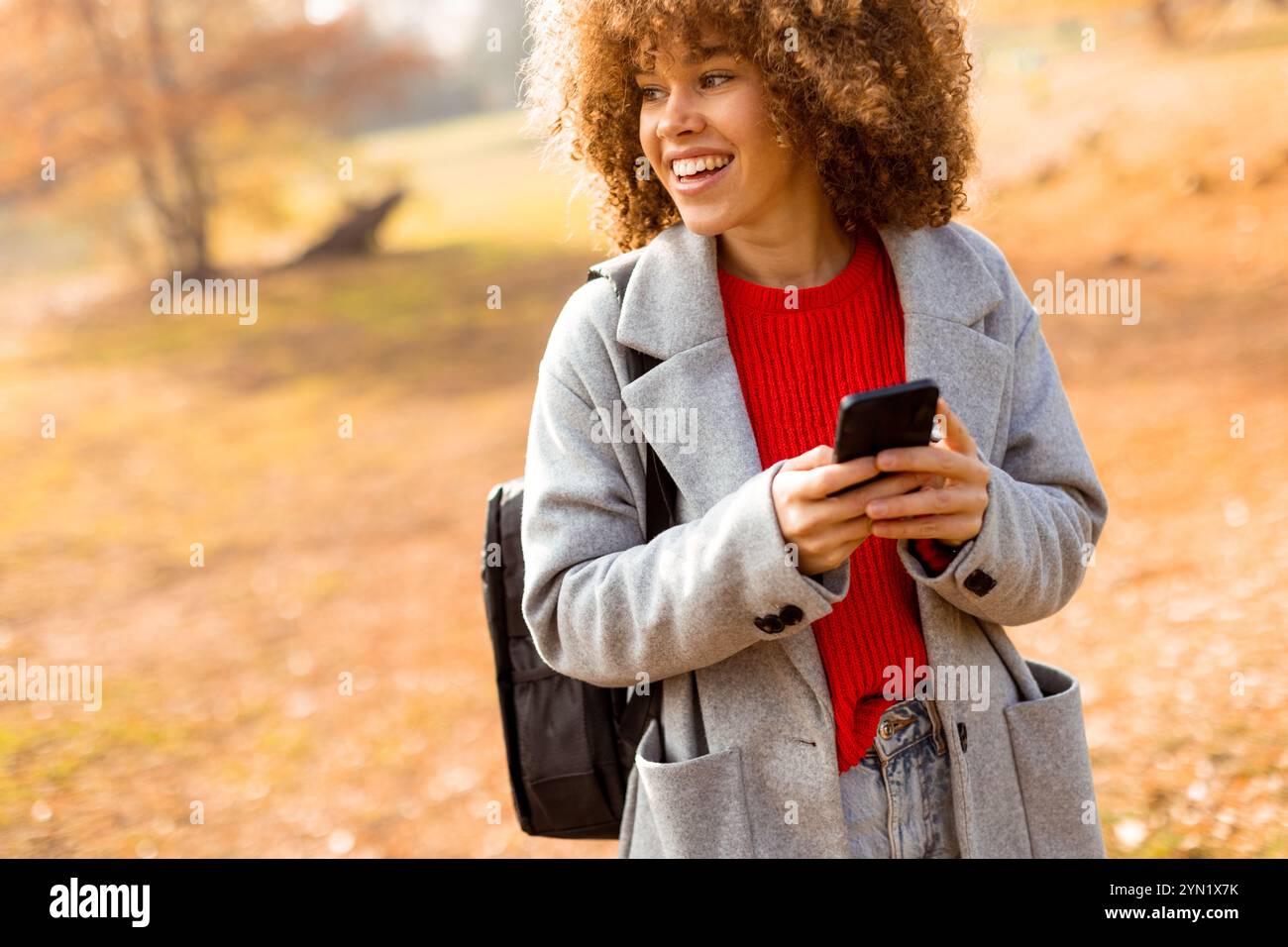 Une personne joyeuse aux cheveux bouclés sourit avec éclat lorsqu'elle s'engage avec son smartphone, portant un pull rouge confortable et un manteau gris élégant au milieu de ses déplacements Banque D'Images