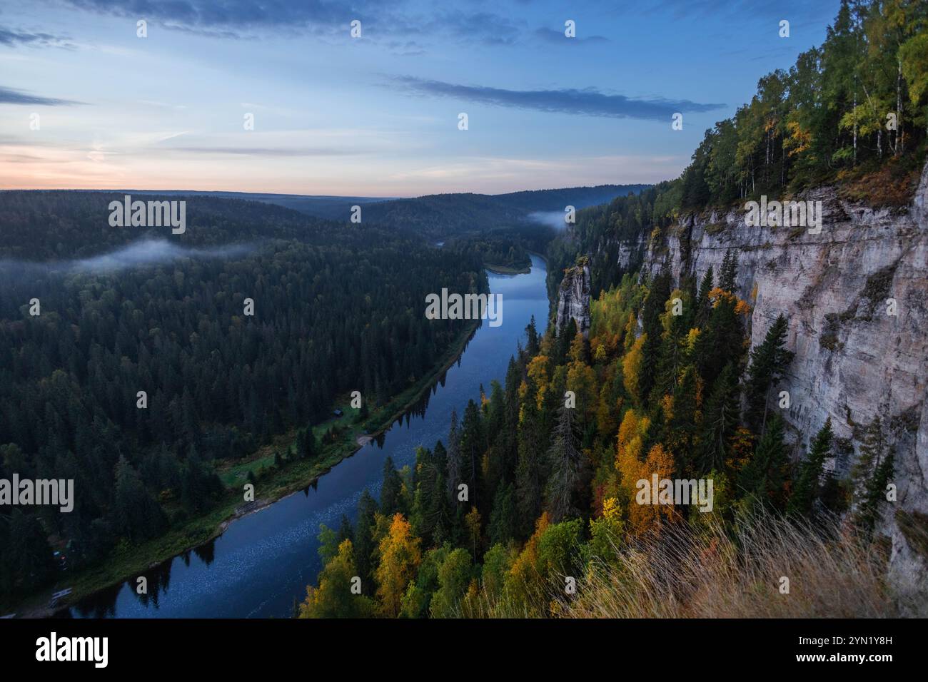 Une rivière tranquille coule doucement à travers des forêts verdoyantes et d'imposantes falaises au crépuscule, avec de la brume s'élevant au-dessus de l'eau et un feuillage automnal vibrant illu Banque D'Images