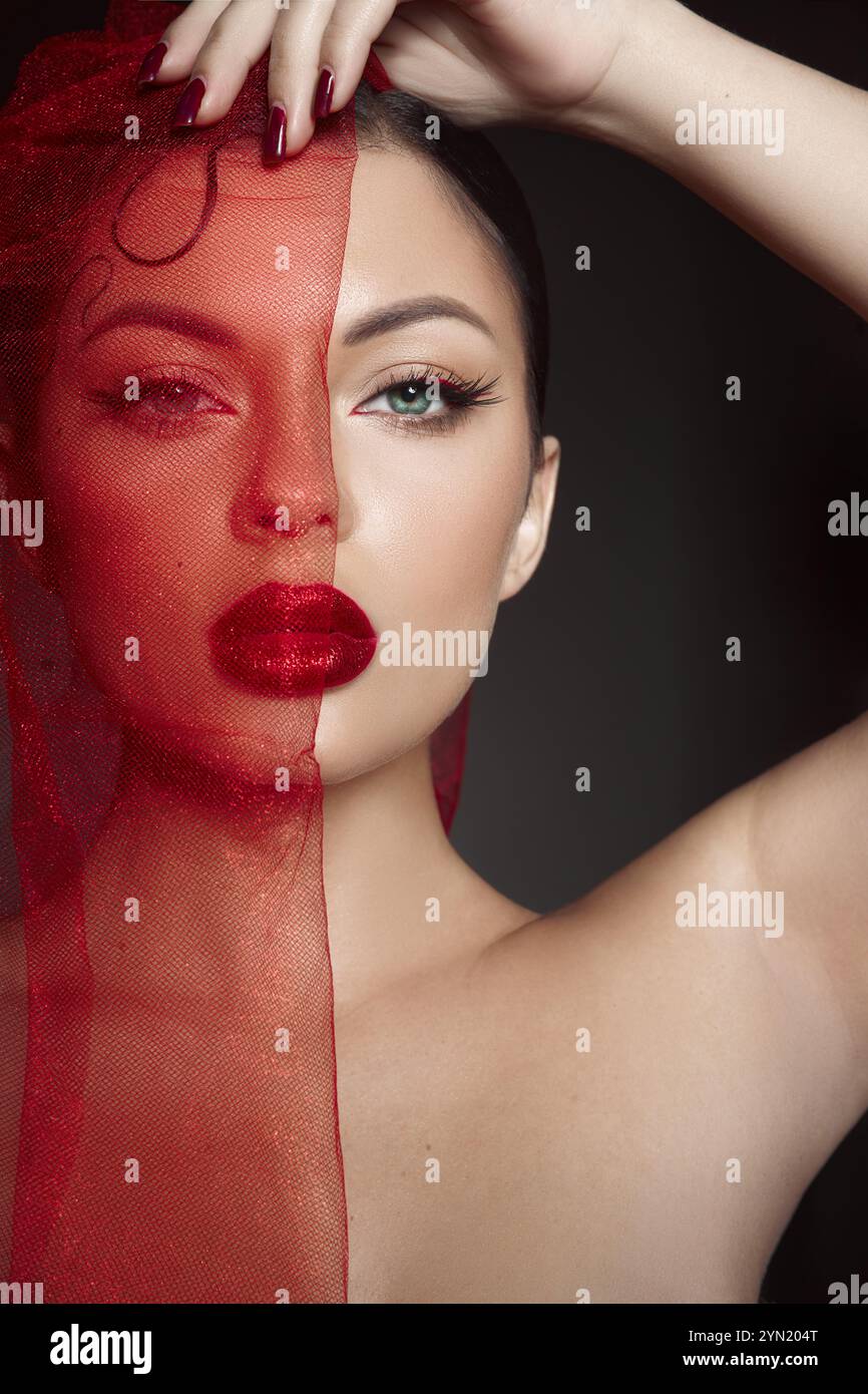 mannequin démontre un look de maquillage artistique en drapant un tissu rouge transparent sur la moitié de son visage, soulignant son maquillage audacieux et ses traits frappants dans un d. Banque D'Images