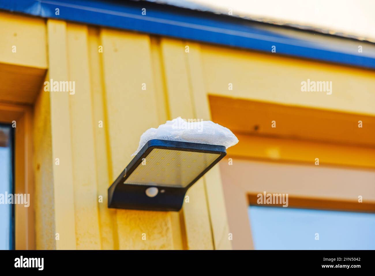Lumière LED extérieure solaire avec capteur de mouvement et neige reposant sur sa surface montée sur une villa murale en bois jaune. Banque D'Images